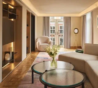 Bright hotel living room with sofa, armchair, round glass tables, and French doors opening to a balcony.