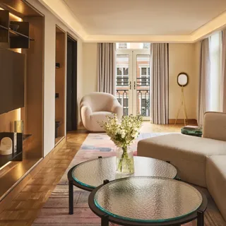 Bright hotel living room with sofa, armchair, round glass tables, and French doors opening to a balcony.