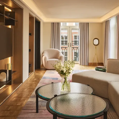 Bright hotel living room with sofa, armchair, round glass tables, and French doors opening to a balcony.