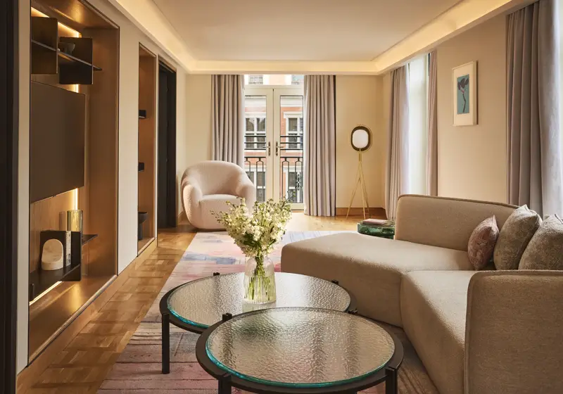 Bright hotel living room with sofa, armchair, round glass tables, and French doors opening to a balcony.