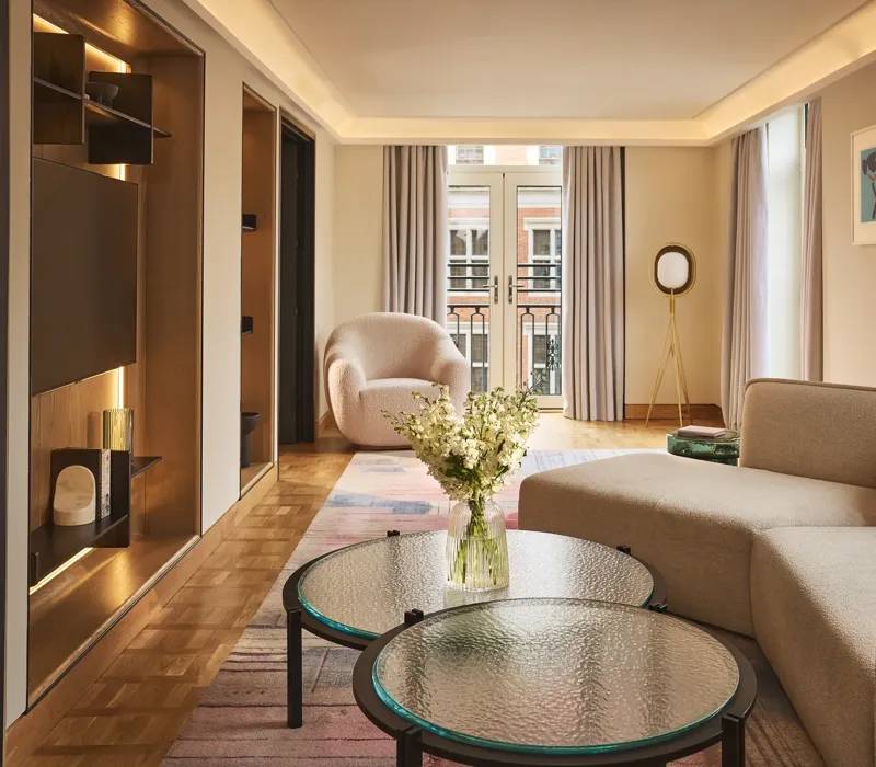 Bright hotel living room with sofa, armchair, round glass tables, and French doors opening to a balcony.