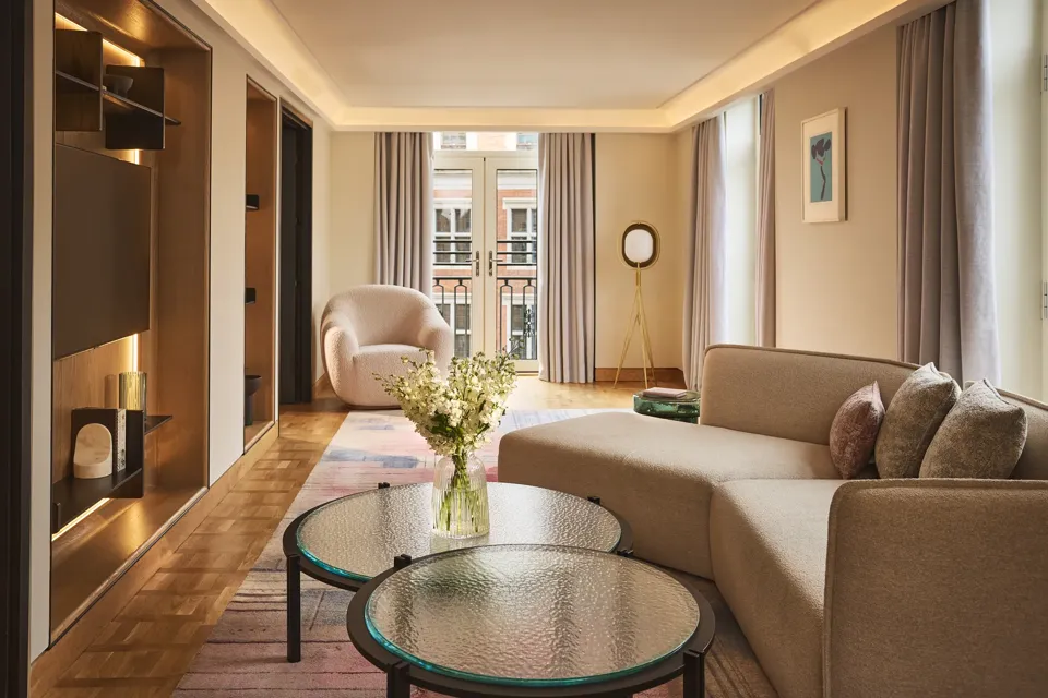 Bright hotel living room with sofa, armchair, round glass tables, and French doors opening to a balcony.