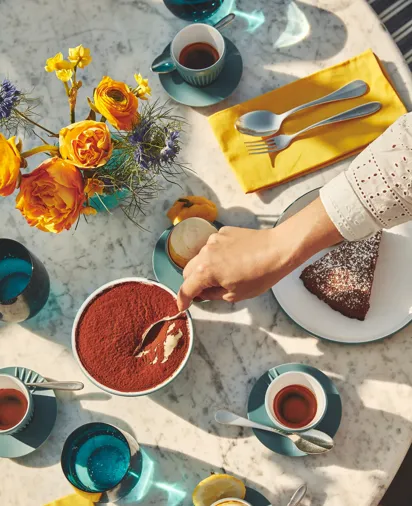 Overhead view of marble table with espresso, cake, tiramisu, and yellow flowers in bright sunlight.