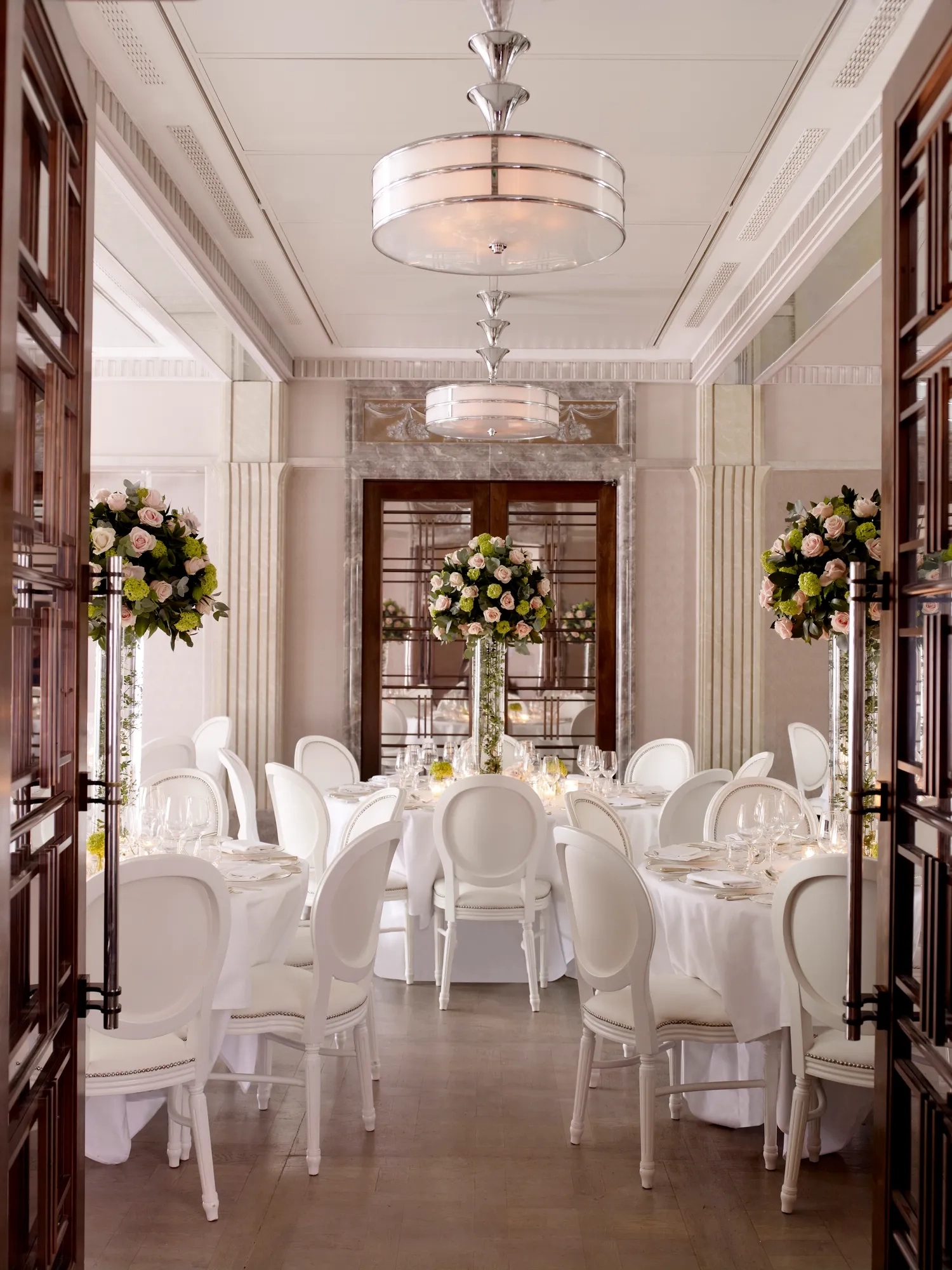 Elegant dining room set with round tables, white chairs, and tall floral centerpieces in soft, natural light.