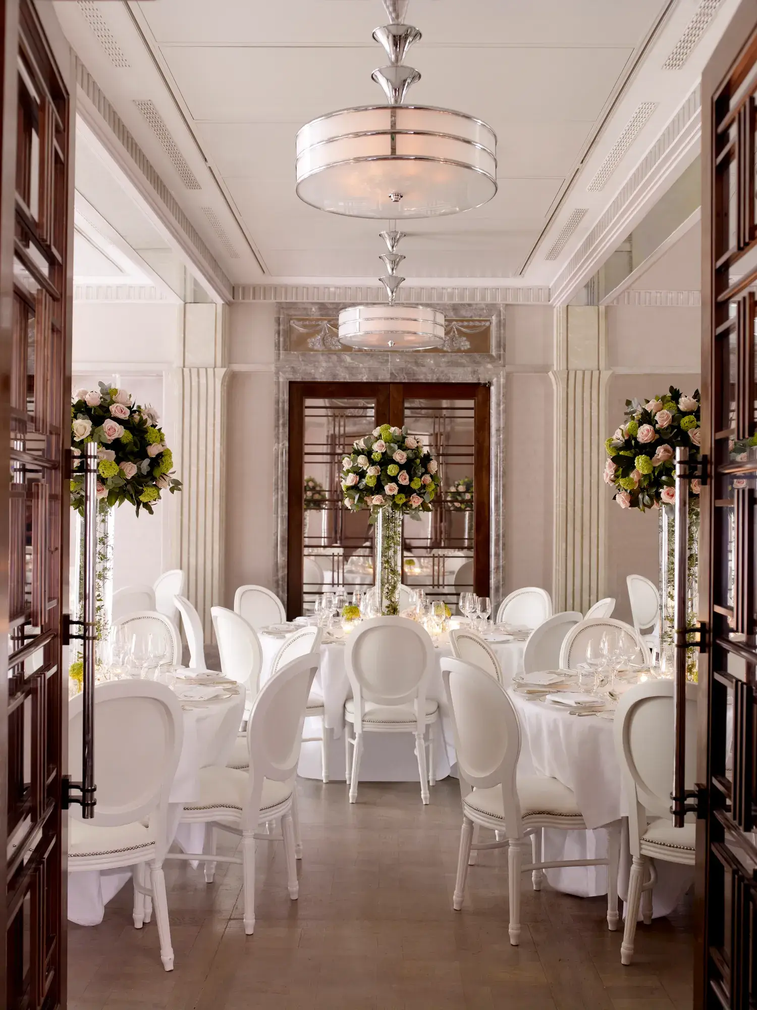 Elegant dining room set with round tables, white chairs, and tall floral centerpieces in soft, natural light.