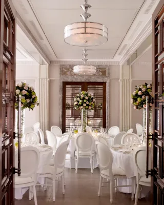 Elegant dining room set with round tables, white chairs, and tall floral centerpieces in soft, natural light.