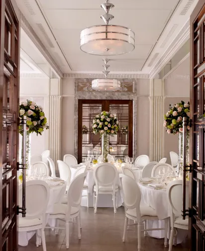Elegant dining room set with round tables, white chairs, and tall floral centerpieces in soft, natural light.