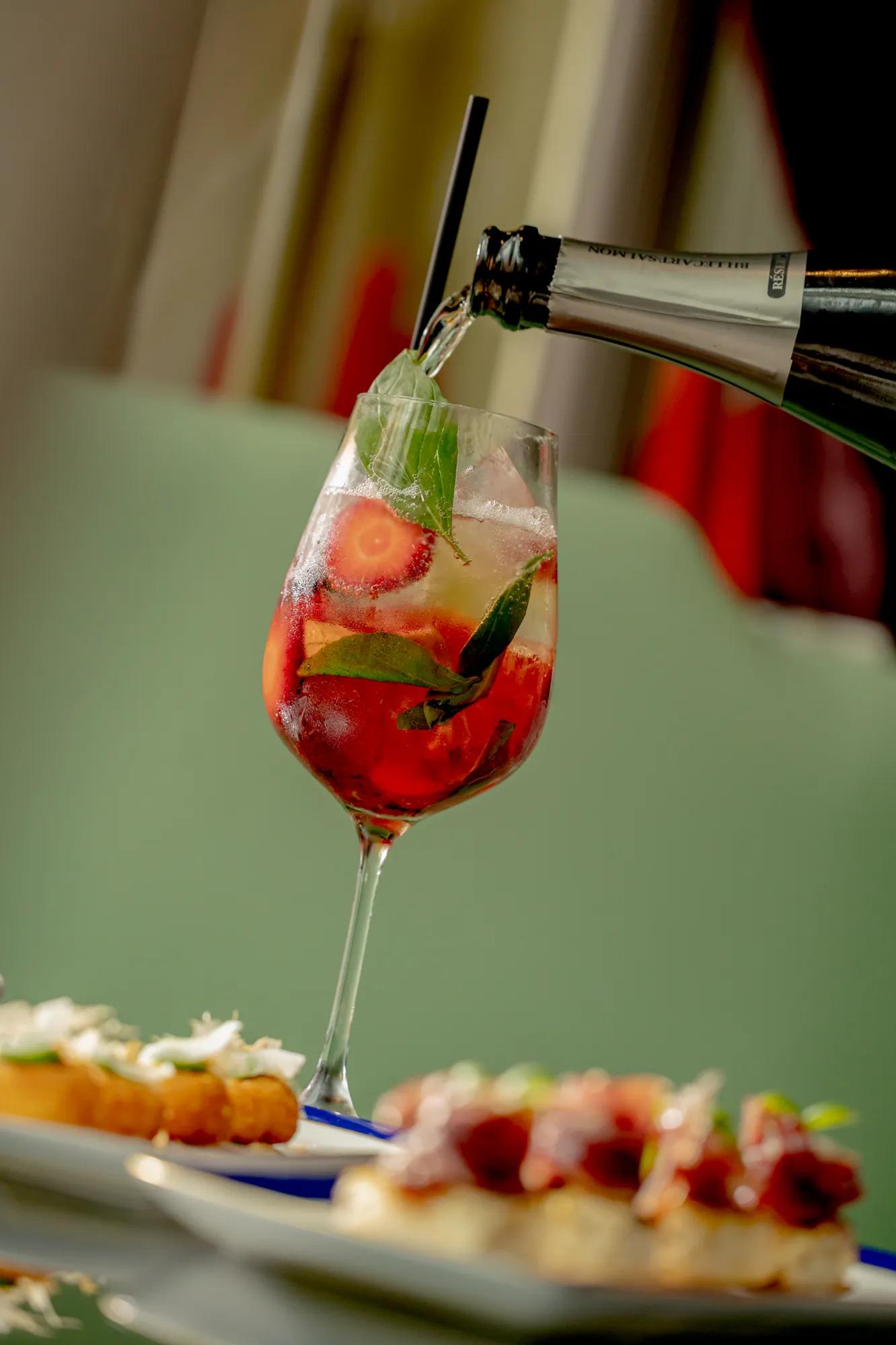 Sparkling drink with strawberries and mint being poured into a glass beside small plated appetisers.