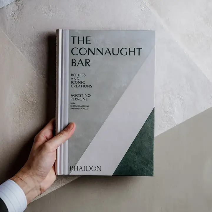 Hand holding “The Connaught Bar” cocktail book against textured neutral wall.