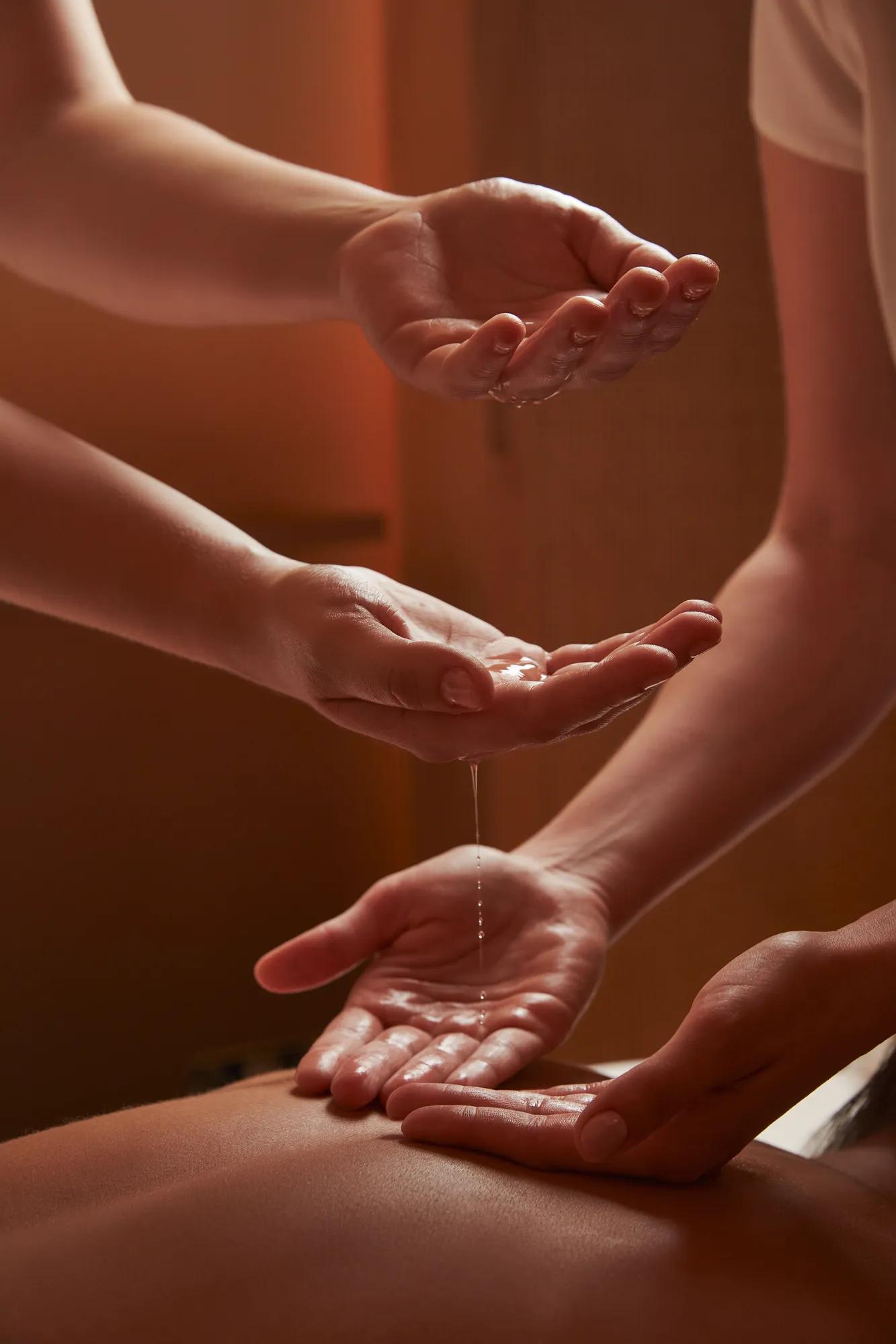 Close-up of two spa therapists pouring warm oil into hands during a treatment in softly lit, tranquil surroundings.