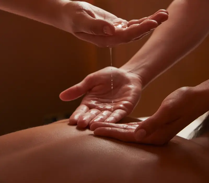 Close-up of two spa therapists pouring warm oil into hands during a treatment in softly lit, tranquil surroundings.
