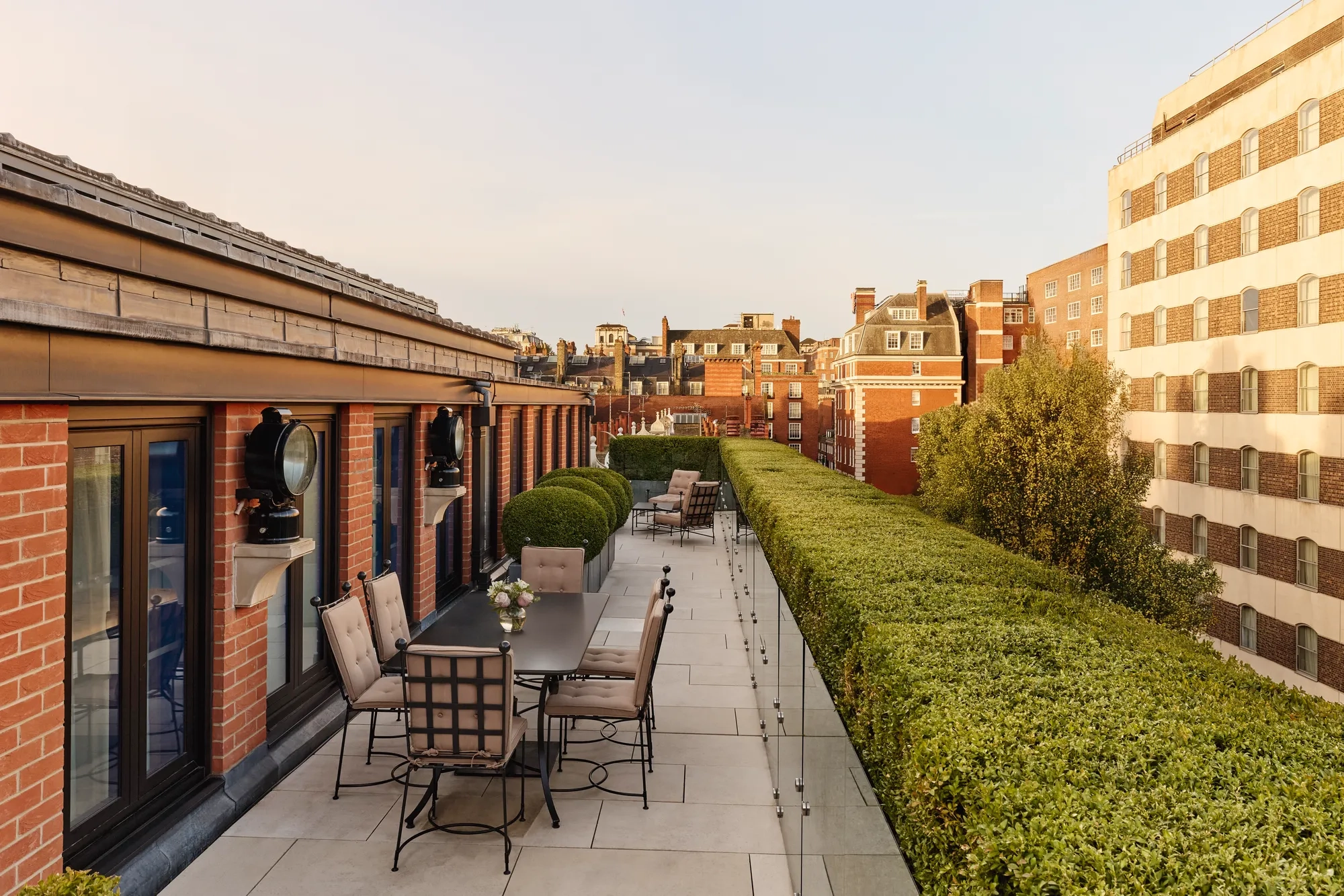 A spacious rooftop terrace with outdoor dining tables and cushioned chairs, bordered by manicured hedges and overlooking the red-brick rooftops and cityscape of Mayfair under warm evening light.