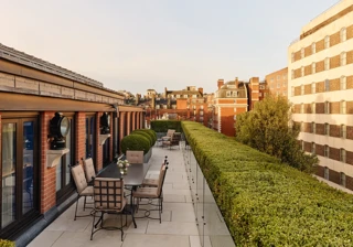 A spacious rooftop terrace with outdoor dining tables and cushioned chairs, bordered by manicured hedges and overlooking the red-brick rooftops and cityscape of Mayfair under warm evening light.