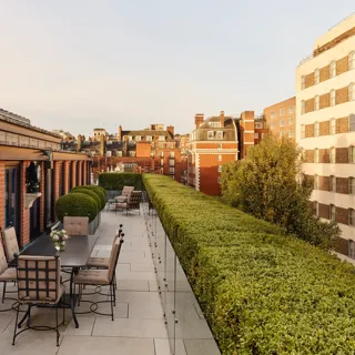 A spacious rooftop terrace with outdoor dining tables and cushioned chairs, bordered by manicured hedges and overlooking the red-brick rooftops and cityscape of Mayfair under warm evening light.