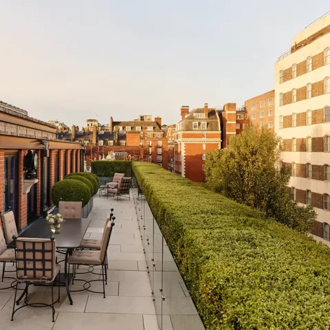 A spacious rooftop terrace with outdoor dining tables and cushioned chairs, bordered by manicured hedges and overlooking the red-brick rooftops and cityscape of Mayfair under warm evening light.