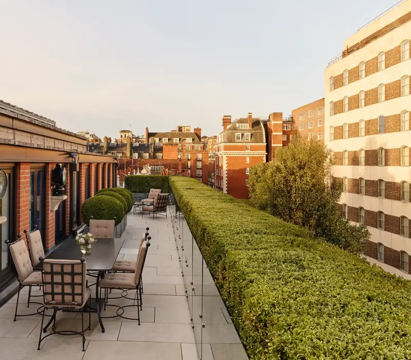 A spacious rooftop terrace with outdoor dining tables and cushioned chairs, bordered by manicured hedges and overlooking the red-brick rooftops and cityscape of Mayfair under warm evening light.