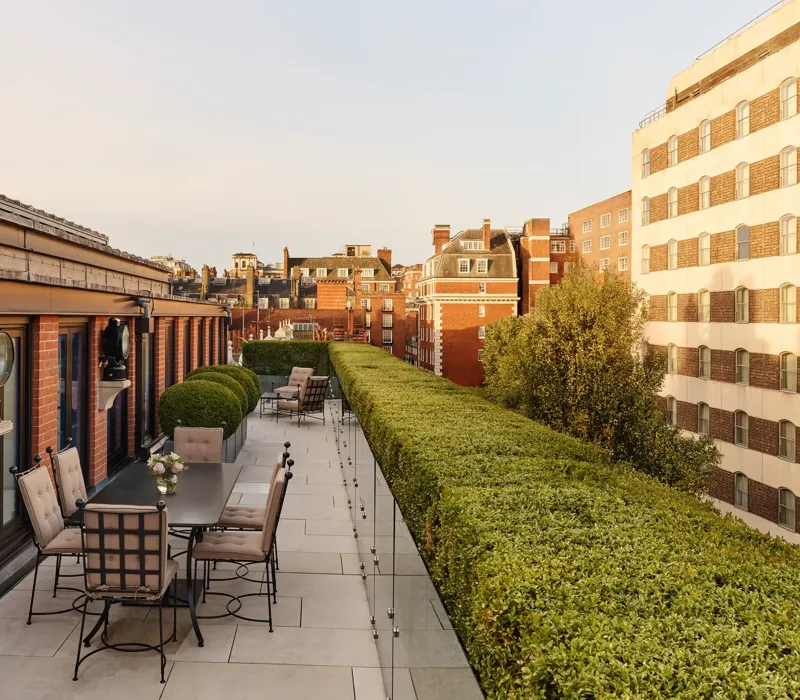 A spacious rooftop terrace with outdoor dining tables and cushioned chairs, bordered by manicured hedges and overlooking the red-brick rooftops and cityscape of Mayfair under warm evening light.
