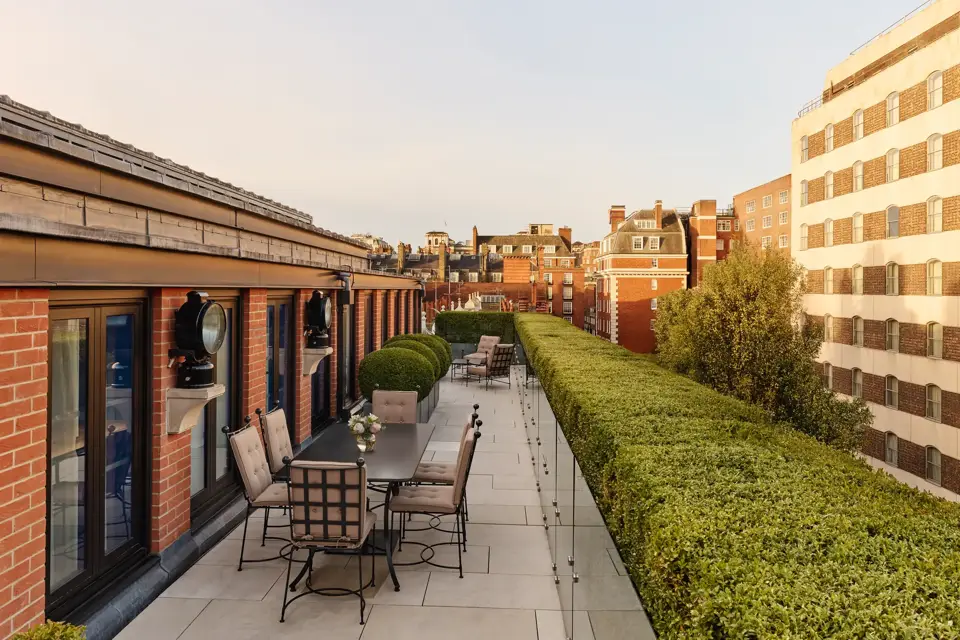 A spacious rooftop terrace with outdoor dining tables and cushioned chairs, bordered by manicured hedges and overlooking the red-brick rooftops and cityscape of Mayfair under warm evening light.