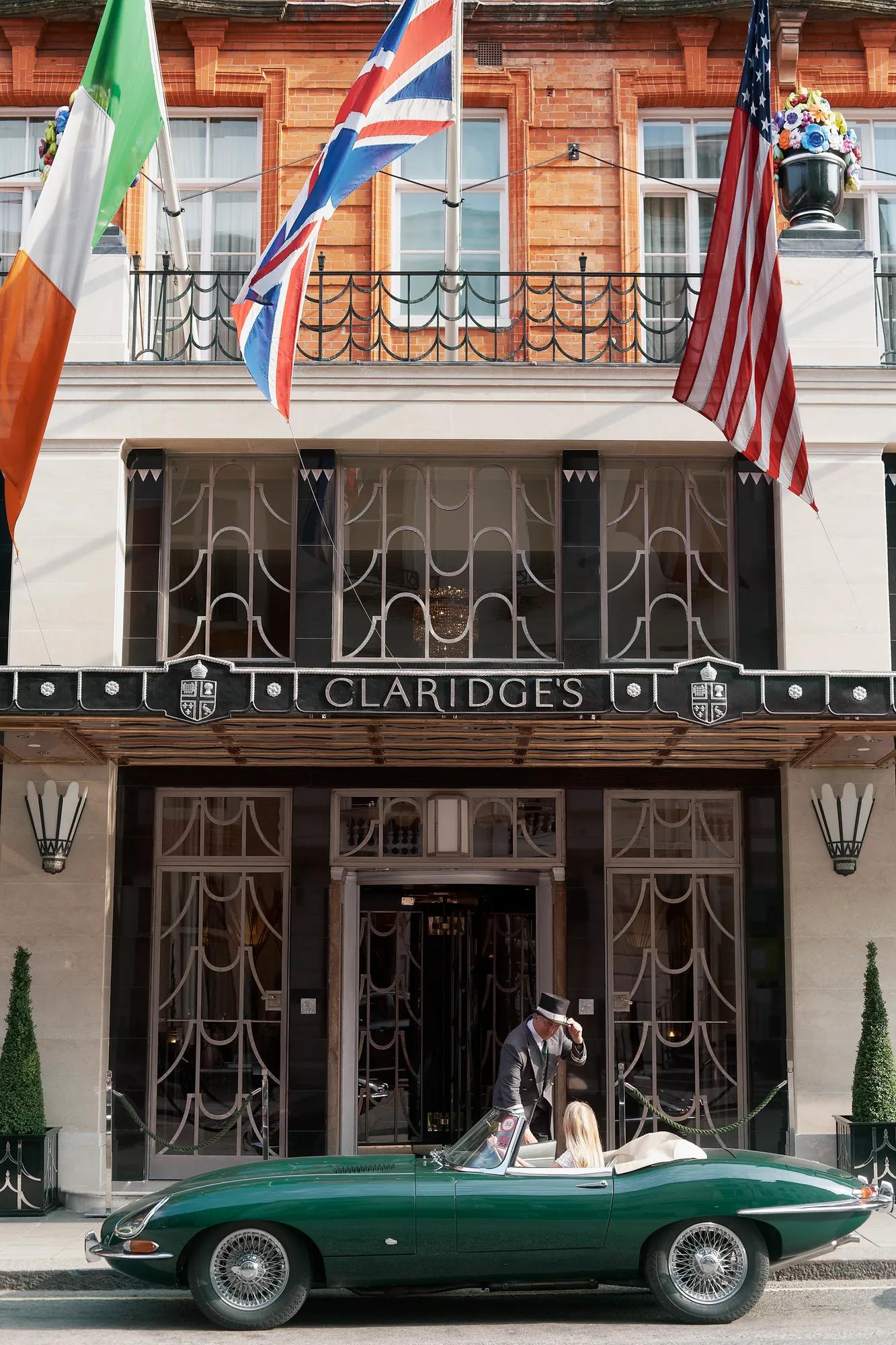 Doorman greeting a guest in a green convertible outside Claridge’s entrance beneath international flags.
