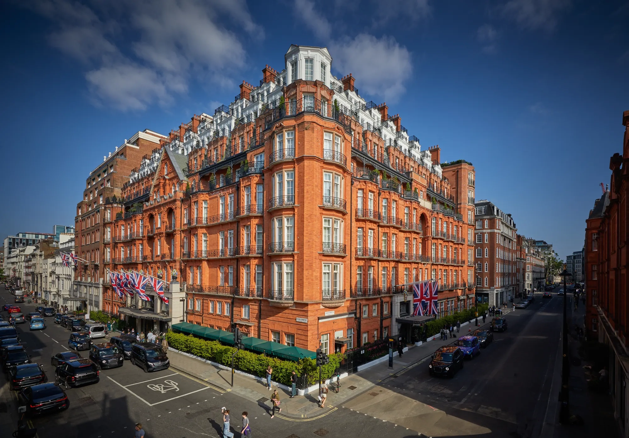 An exterior view of Claridge’s hotel in Mayfair, London, showing its grand red-brick façade adorned with Union Jack flags, green awnings, and black taxis along the street under a bright blue sky.