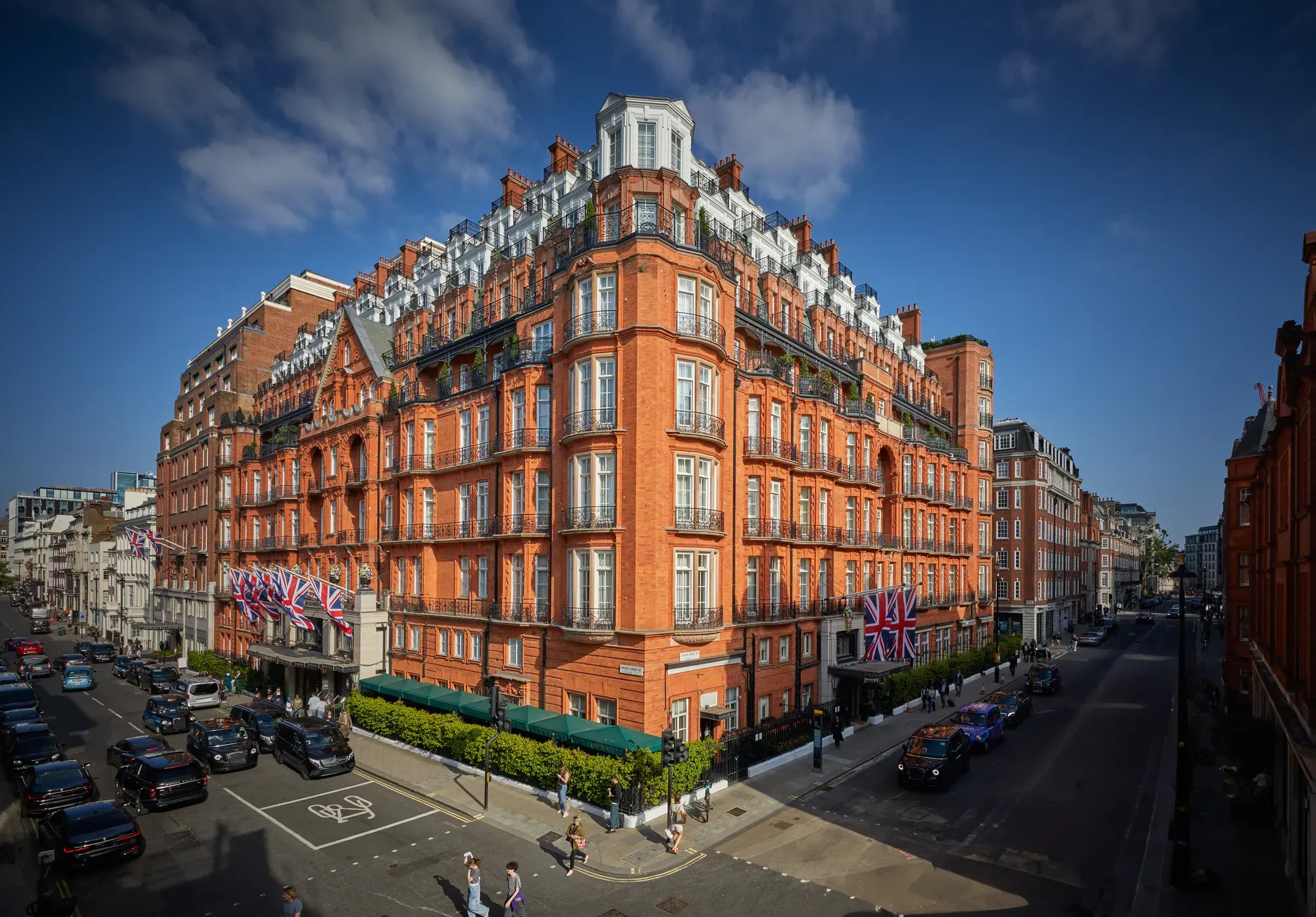 An exterior view of Claridge’s hotel in Mayfair, London, showing its grand red-brick façade adorned with Union Jack flags, green awnings, and black taxis along the street under a bright blue sky.