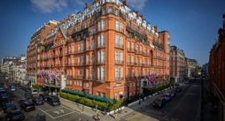 An exterior view of Claridge’s hotel in Mayfair, London, showing its grand red-brick façade adorned with Union Jack flags, green awnings, and black taxis along the street under a bright blue sky.