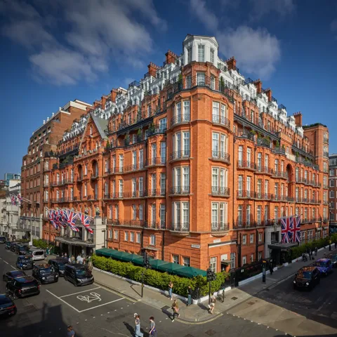 An exterior view of Claridge’s hotel in Mayfair, London, showing its grand red-brick façade adorned with Union Jack flags, green awnings, and black taxis along the street under a bright blue sky.