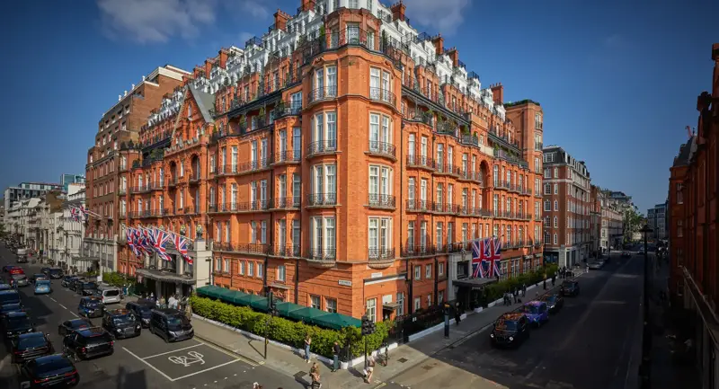 An exterior view of Claridge’s hotel in Mayfair, London, showing its grand red-brick façade adorned with Union Jack flags, green awnings, and black taxis along the street under a bright blue sky.