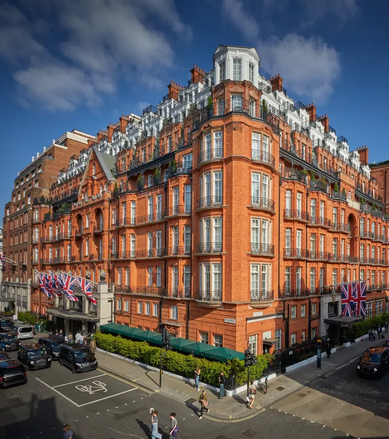 An exterior view of Claridge’s hotel in Mayfair, London, showing its grand red-brick façade adorned with Union Jack flags, green awnings, and black taxis along the street under a bright blue sky.