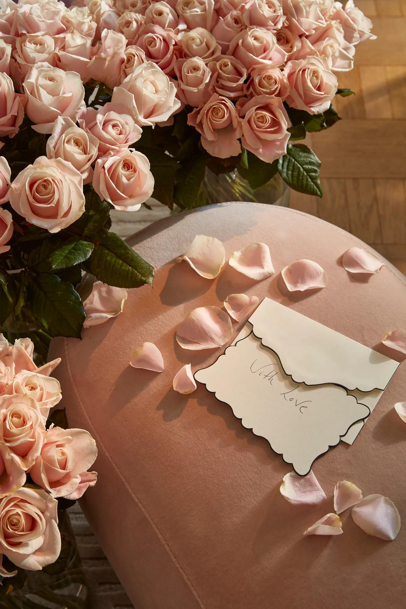 Close-up of pale pink roses and scattered petals on a soft pink velvet surface, with an envelope reading “With love” placed among the flowers.