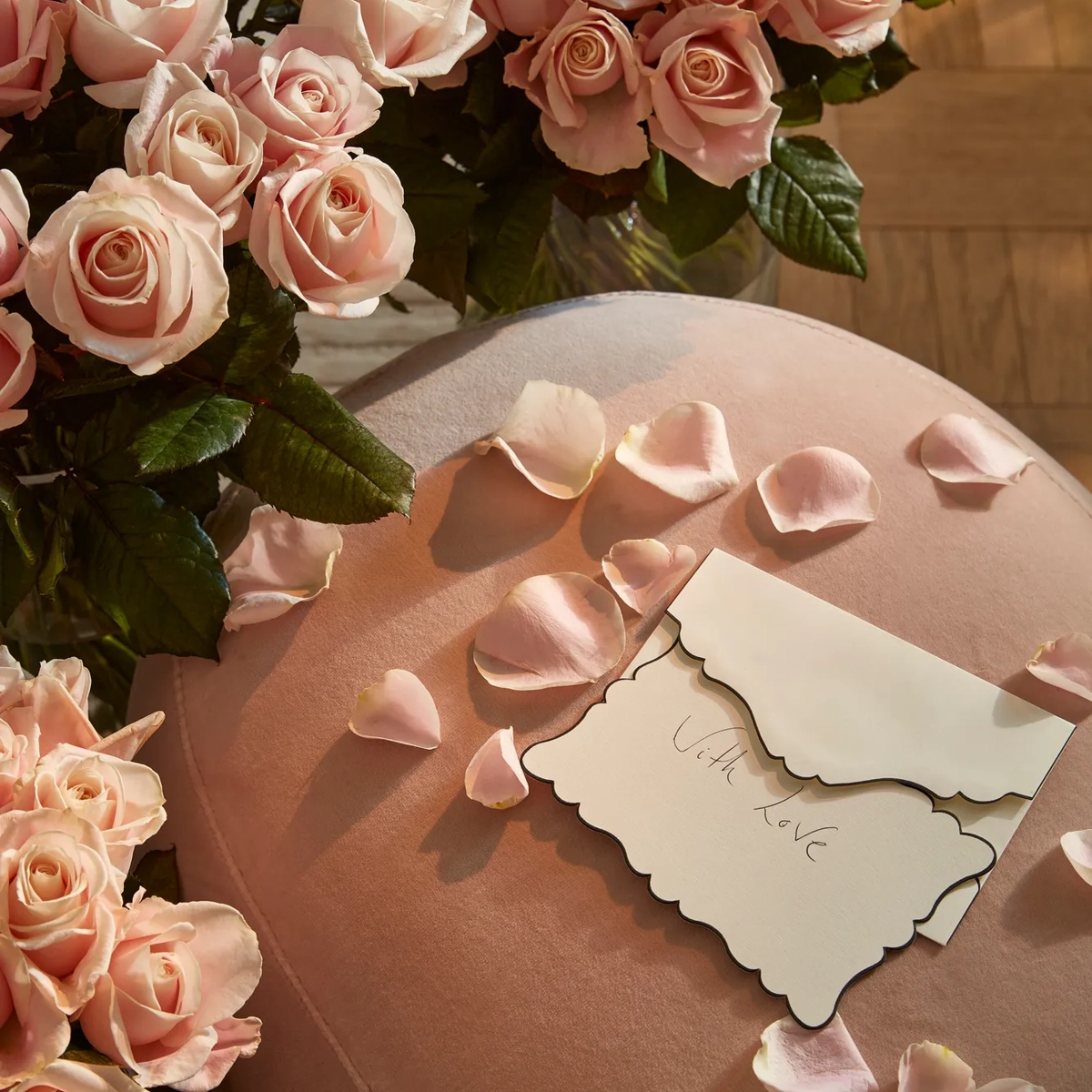 Close-up of pale pink roses and scattered petals on a soft pink velvet surface, with an envelope reading “With love” placed among the flowers.