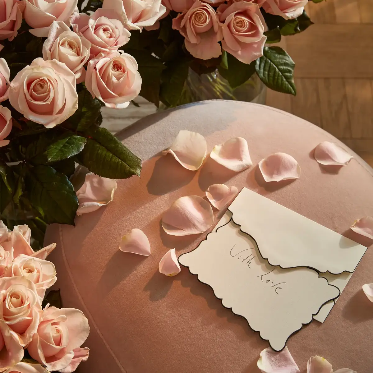 Close-up of pale pink roses and scattered petals on a soft pink velvet surface, with an envelope reading “With love” placed among the flowers.
