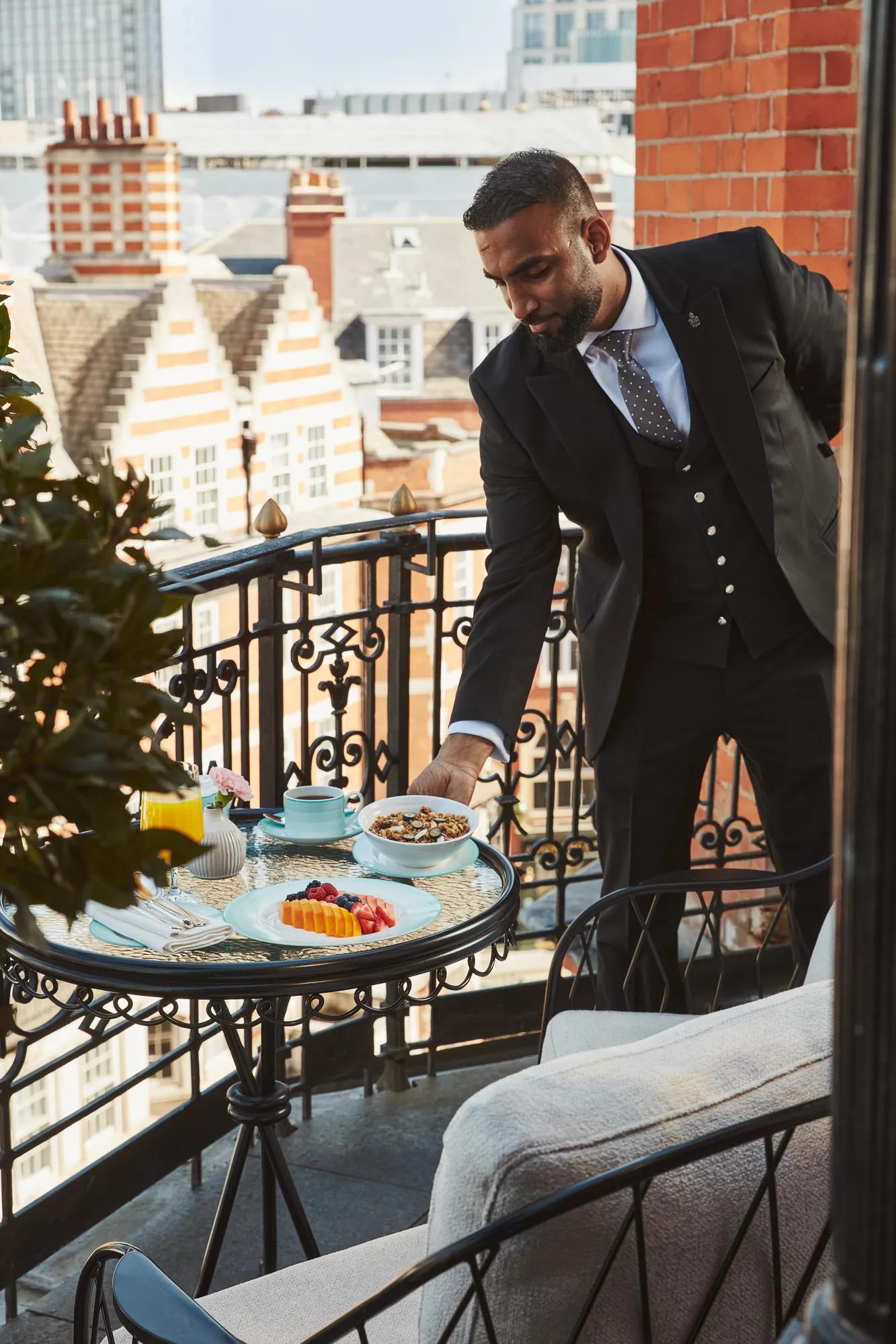 Hotel staff member in a suit setting a breakfast table with fruit and pastries on a private balcony overlooking Mayfair rooftops.