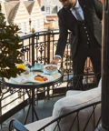 Hotel staff member in a suit setting a breakfast table with fruit and pastries on a private balcony overlooking Mayfair rooftops.