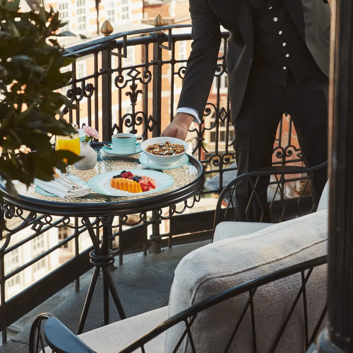 Hotel staff member in a suit setting a breakfast table with fruit and pastries on a private balcony overlooking Mayfair rooftops.