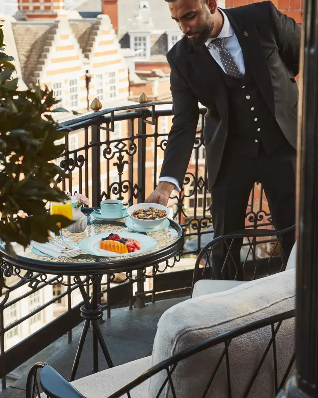 Hotel staff member in a suit setting a breakfast table with fruit and pastries on a private balcony overlooking Mayfair rooftops.