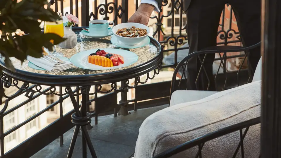 Hotel staff member in a suit setting a breakfast table with fruit and pastries on a private balcony overlooking Mayfair rooftops.