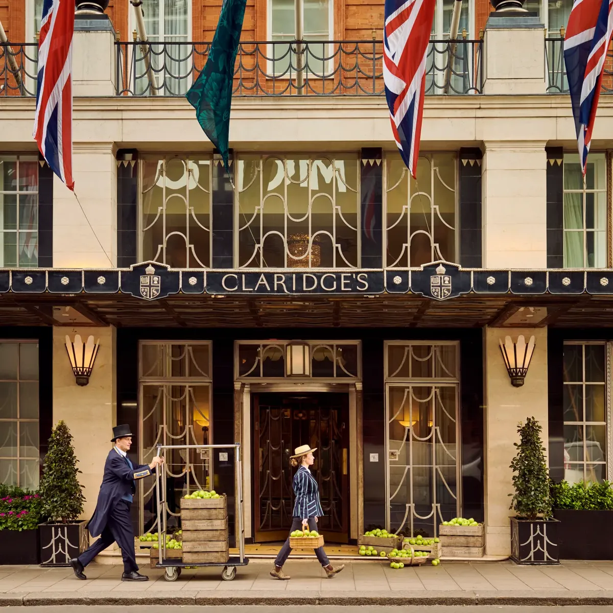 Claridge’s hotel entrance with Union Jack flags, a doorman pushing luggage trolley, and a guest walking past.