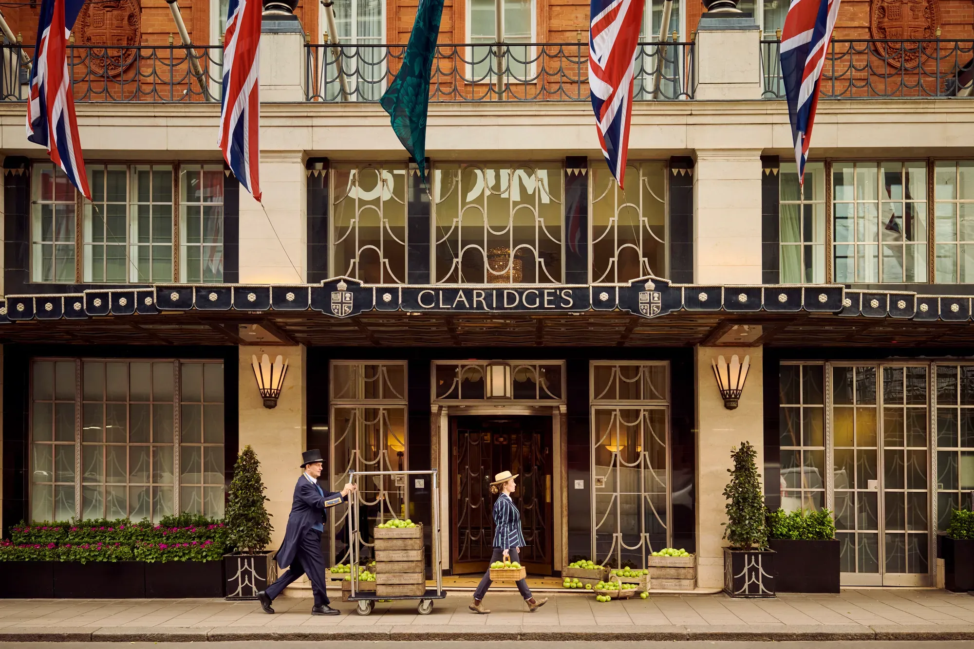 Claridge’s hotel entrance with Union Jack flags, a doorman pushing luggage trolley, and a guest walking past.