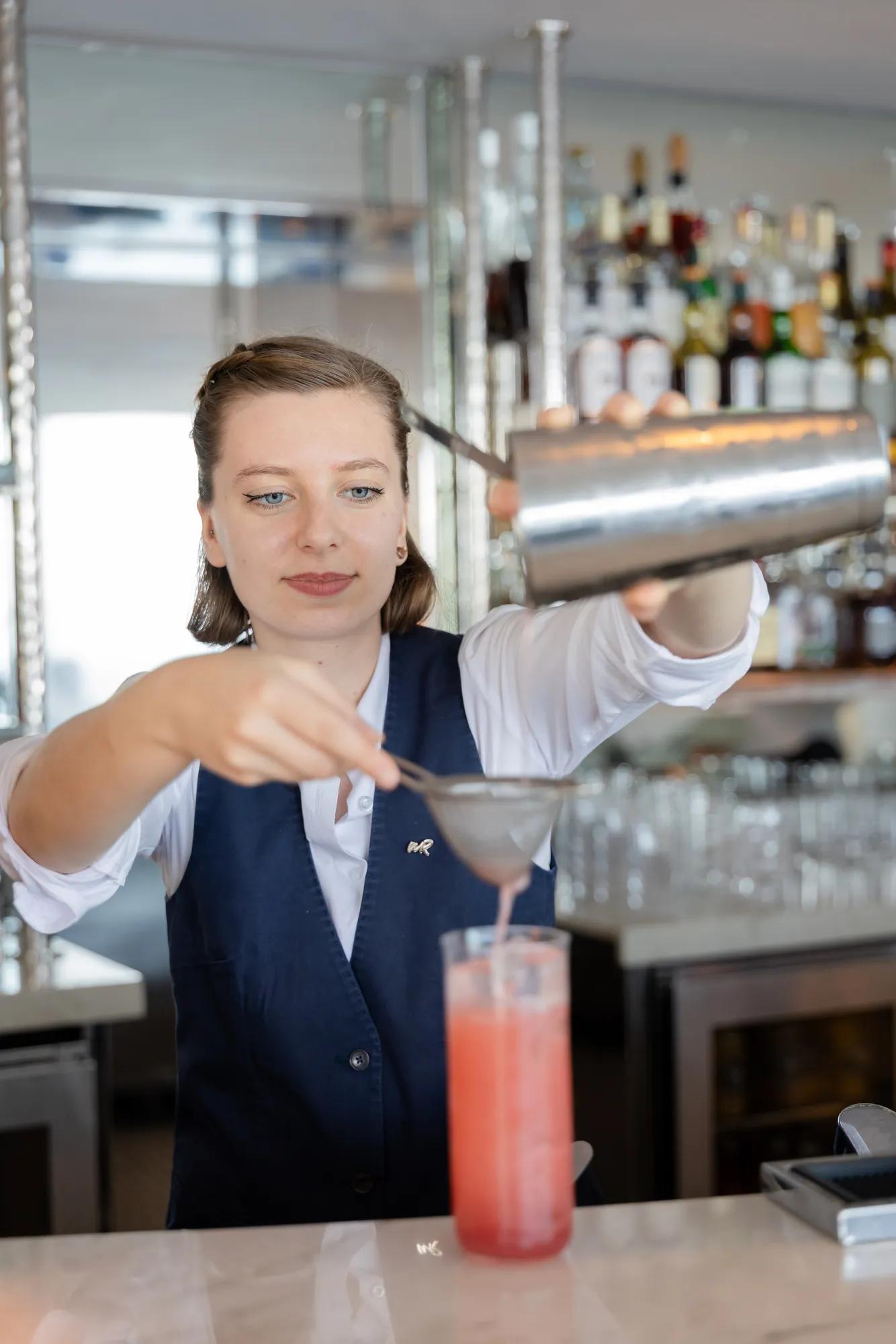 Bartender straining a pink cocktail into a tall glass behind a well-stocked bar with bottles and glassware.