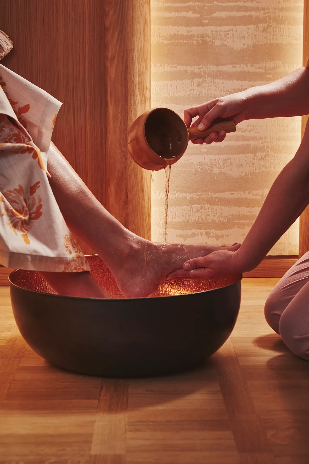 A spa therapist pours warm water over a guest’s feet during a foot ritual, with gentle lighting and wooden surroundings at The Maybourne Riviera.