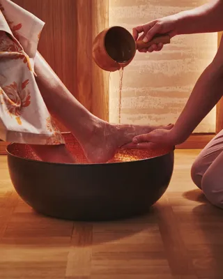 A spa therapist pours warm water over a guest’s feet during a foot ritual, with gentle lighting and wooden surroundings at The Maybourne Riviera.