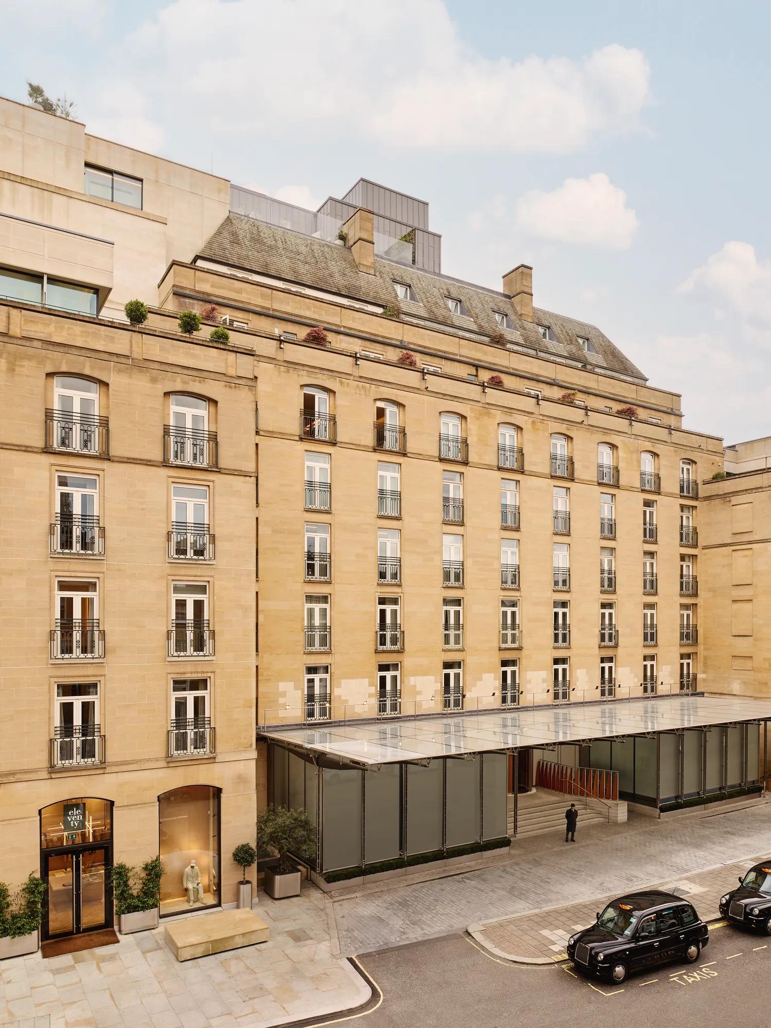 Stone hotel façade with rows of French windows and balconies, a glass entrance canopy, and black taxis on the street outside.