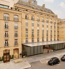 Stone hotel façade with rows of French windows and balconies, a glass entrance canopy, and black taxis on the street outside.