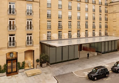 Stone hotel façade with rows of French windows and balconies, a glass entrance canopy, and black taxis on the street outside.
