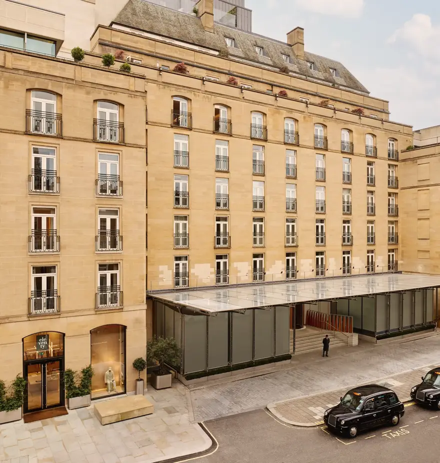 Stone hotel façade with rows of French windows and balconies, a glass entrance canopy, and black taxis on the street outside.