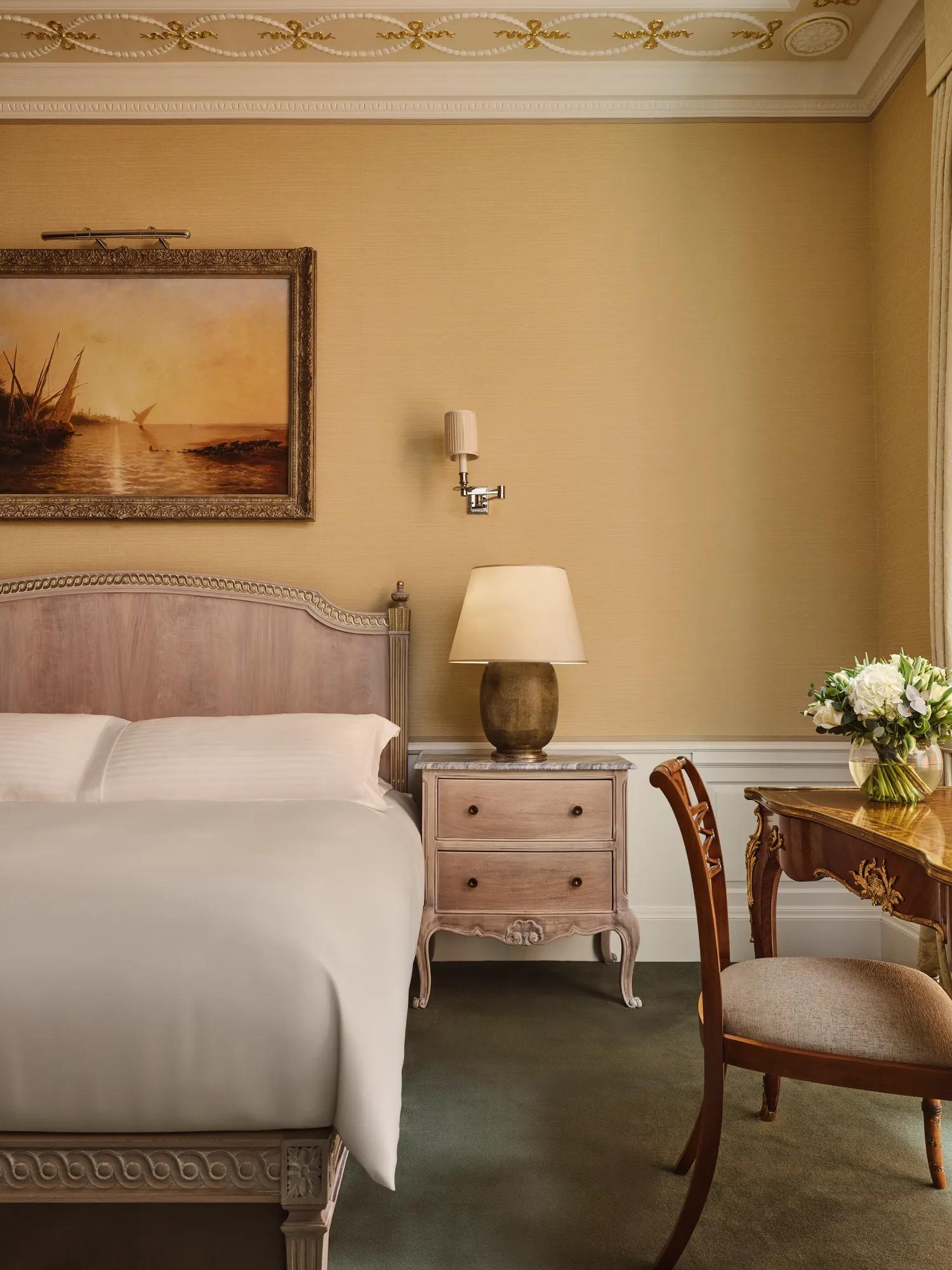 Classic bedroom with upholstered bed, bedside table and lamp, framed landscape painting, and writing desk with flowers beside tall window.