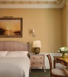 Classic bedroom with upholstered bed, bedside table and lamp, framed landscape painting, and writing desk with flowers beside tall window.