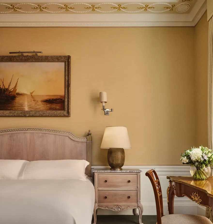 Classic bedroom with upholstered bed, bedside table and lamp, framed landscape painting, and writing desk with flowers beside tall window.