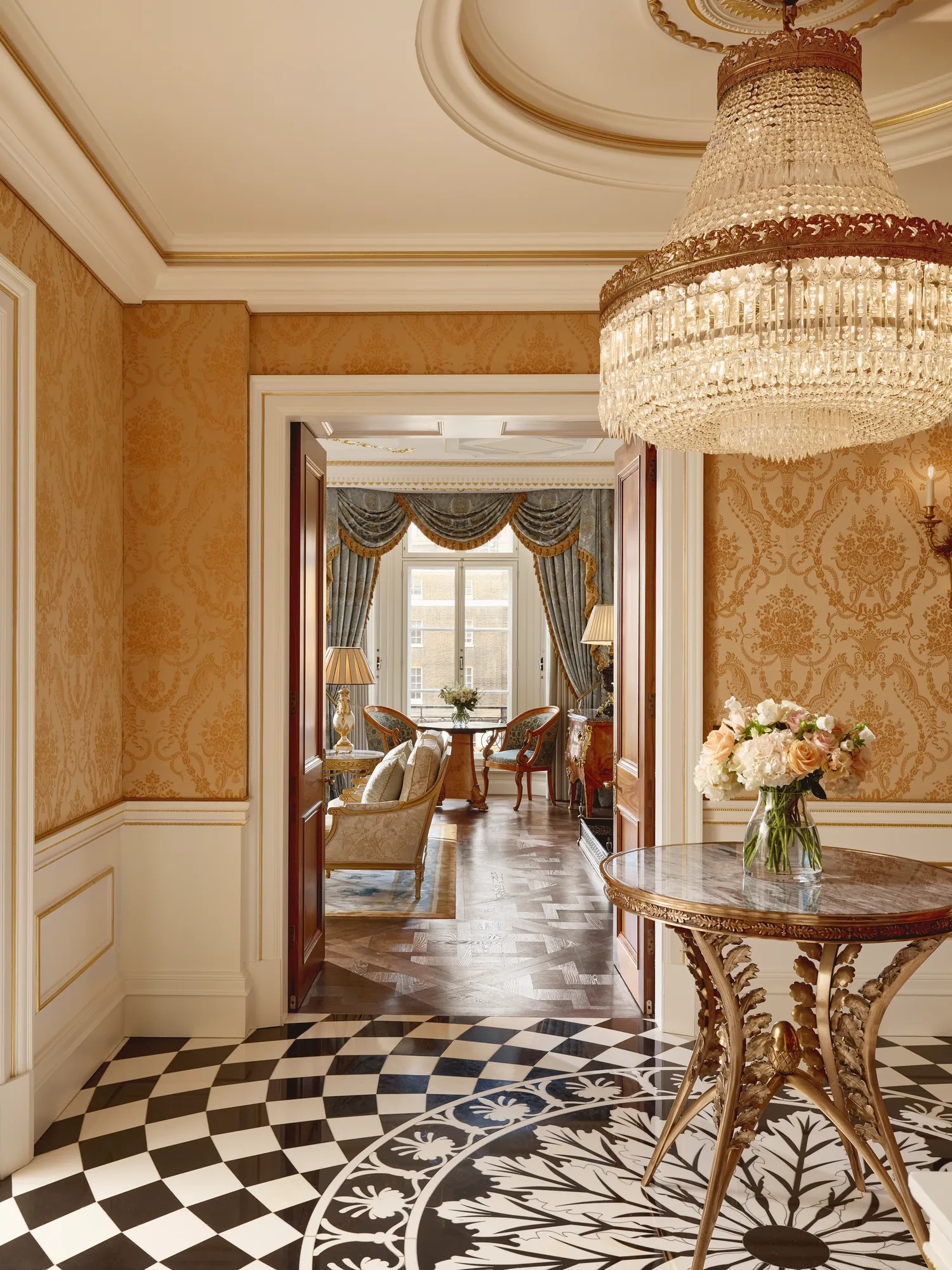 Elegant hallway with gold damask wallpaper, a round marble-topped table holding a vase of flowers, a crystal chandelier, and a view into a sitting room with blue drapes and classic furnishings.